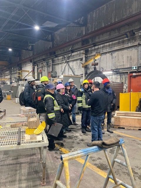 Travailleurs en tenue de sécurité et casques colorés écoutant un superviseur dans une usine, entourés d'équipements industriels et de tables métalliques.
