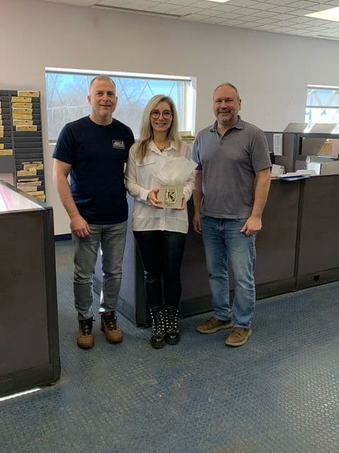 Trois personnes souriantes debout dans un bureau, une femme au centre tenant un cadeau emballé avec un logo en forme de "K".