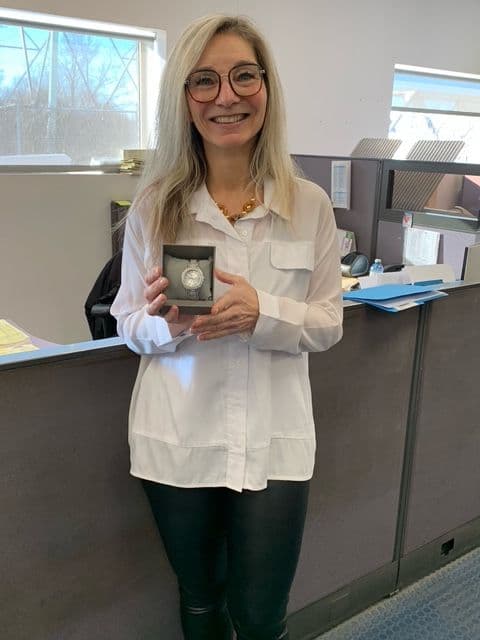 Femme souriante aux cheveux blonds et lunettes, tenant une boîte contenant une montre argentée, debout dans un bureau.